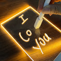 LED glow note board lit warm on a wooden desk, a hand writing a message on it with the white marker pen
