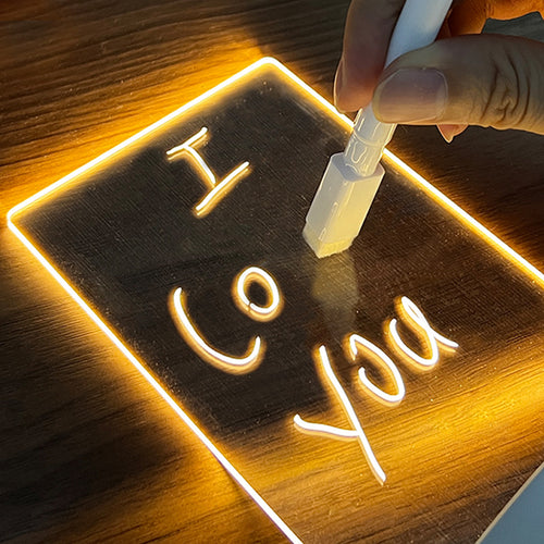 LED glow note board lit warm on a wooden desk, a hand writing a message on it with the white marker pen