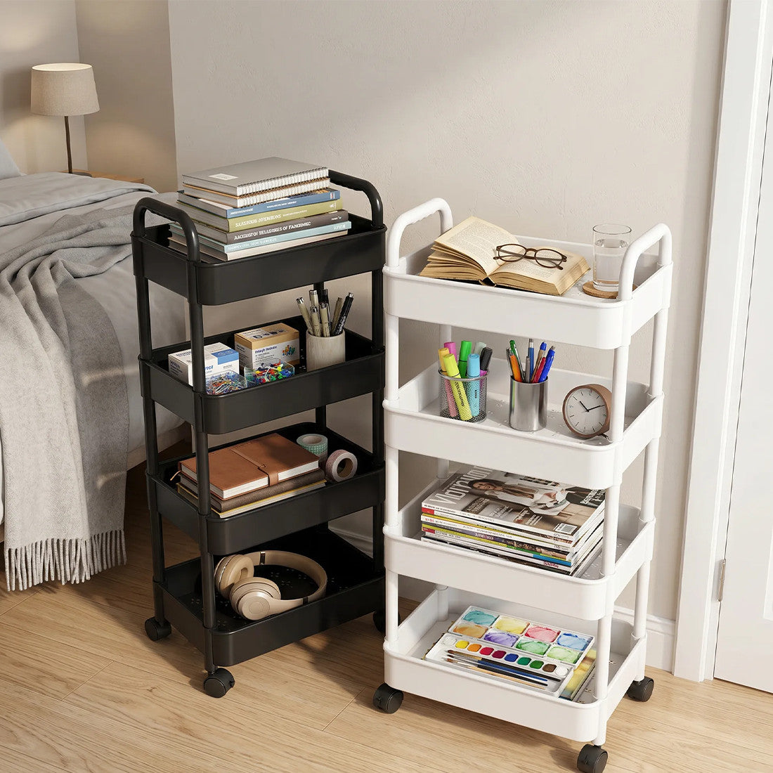 A black and a white four-tier rolling trolley side by side in a bedroom, tiers holding books, stationery and headphones.